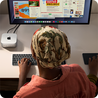 Persona pie rakstāmgalda raksta ar Mac mini, kas savienots ar Studio Display, Magic Keyboard tastatūru un Magic Trackpad skārienpaliktni, apkārt dažādi priekšmeti