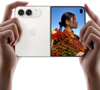 iPhone Duo v nebeskej bielej farbe, držia ho dve ruky v orientácii na šírku, je rozložený, na ľavom paneli vidno 48 Mpx zadné kamery Dual Fusion, na pravom paneli vidno na vonkajšom displeji fotografovanú osobu, ktorá vďaka funkcii Duo Preview vidí pri fotení svoju tvár