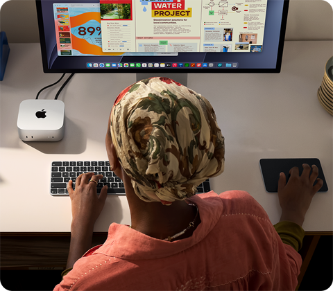 Niekto píše pri stole, na ktorom je Mac mini s pripojeným displejom Studio Display, klávesnica Magic Keyboard, trackpad Magic Trackpad a rôzne predmety
