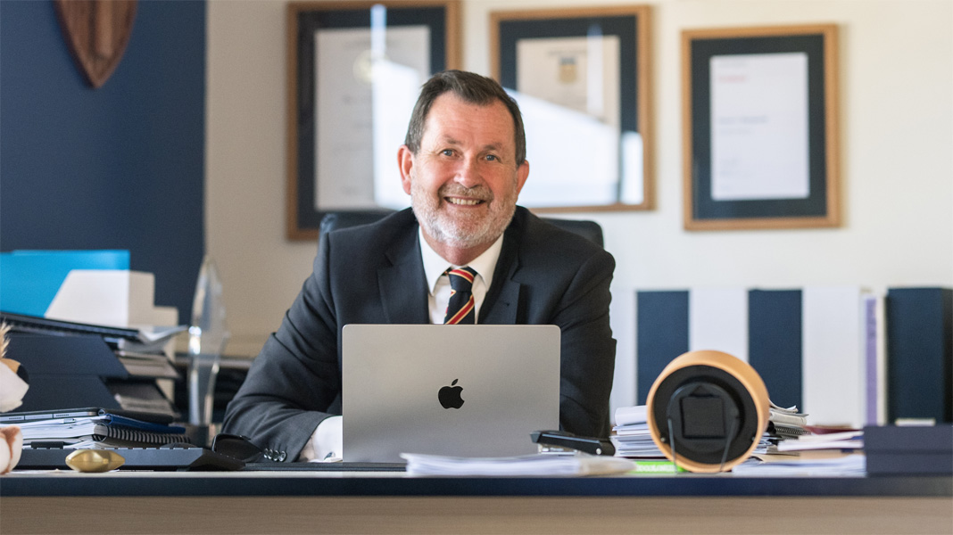 A principal sits at his desk behind an open Apple laptop, smiling at the camera.