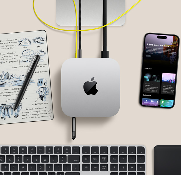 Mac mini, atas, aluminium perak, sudut melengkung, logo Apple hitam mengilap di tengah, terhubung dengan kabel, stik USB dimasukkan ke port USB-C di depan, dudukan Studio Display sebagian terlihat, iPhone, Magic Trackpad, Magic Keyboard dengan Touch ID, stylus dan pad sketsa tinta elektronik