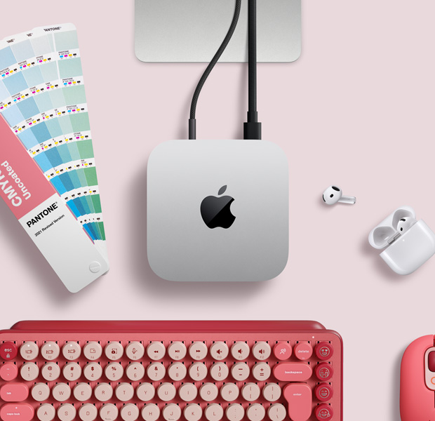Mac mini, atas, aluminium perak, sudut melengkung, logo Apple hitam mengilap di tengah, terhubung dengan kabel, dudukan Studio Display sebagian terlihat, AirPods 4, sampel warna Pantone, serta keyboard dan mouse Logitech merah